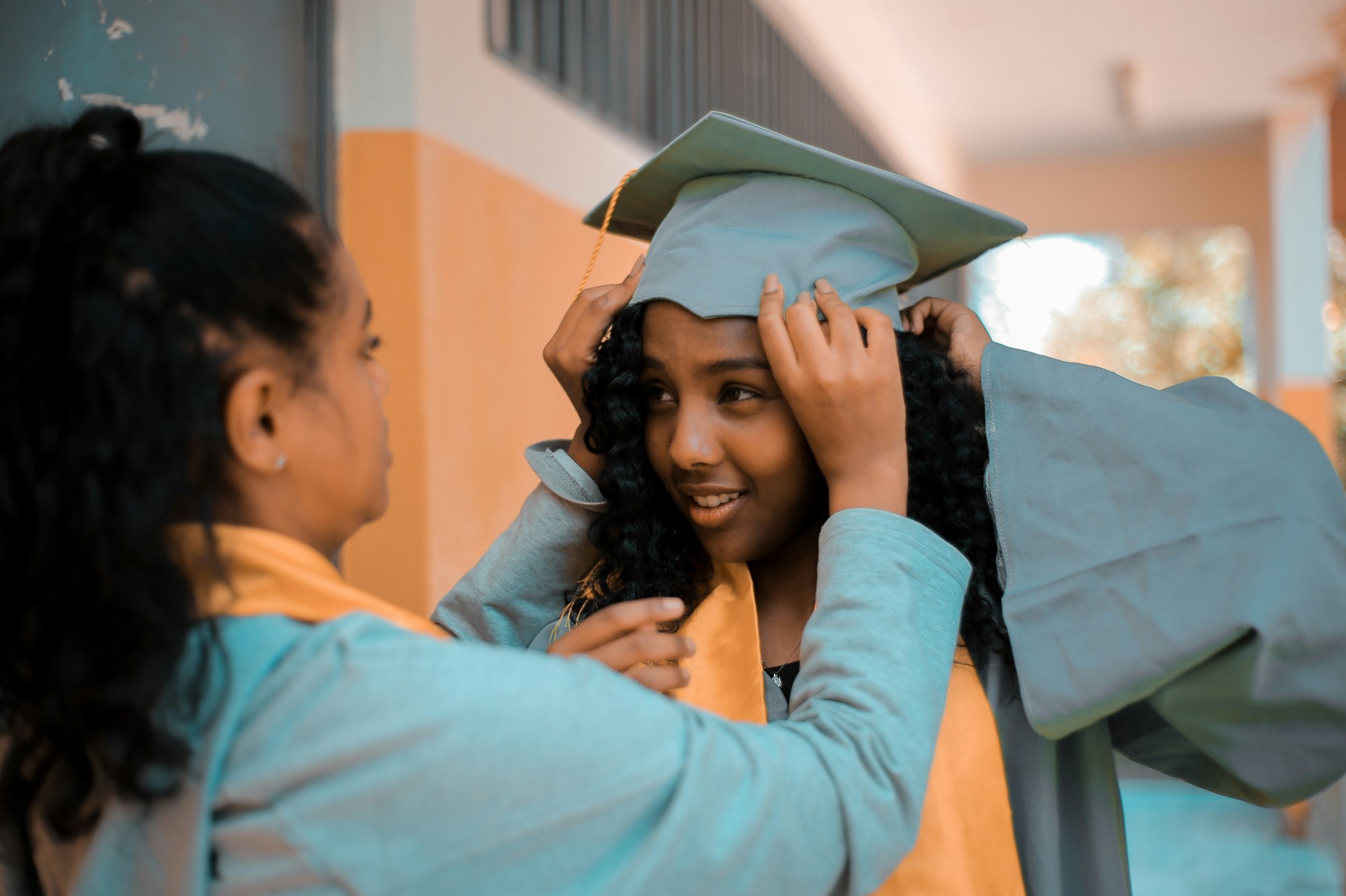 Jovem ajustando o capelo de formatura de outra jovem, as duas sorrindo.